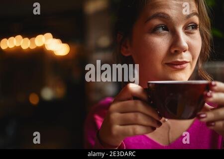 Thoughtful young woman sitting in coffee house and looking out the window while drinking tea Stock Photo