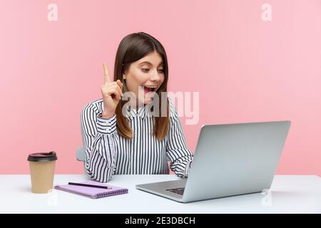 Smiling positive woman office worker in striped shirt pointing finger up talking video call on laptop, great idea for startup, business solution. Indo Stock Photo