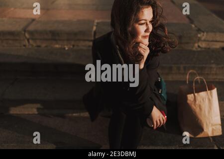 Happy pretty lady sitting on steps and looking away in the city during the evening Stock Photo