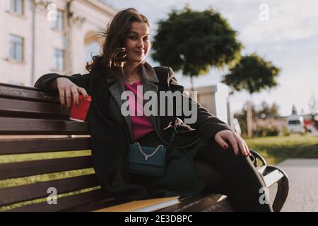 Happy young lady with bag sitting on bench while enjoying the hot drink in the city Stock Photo