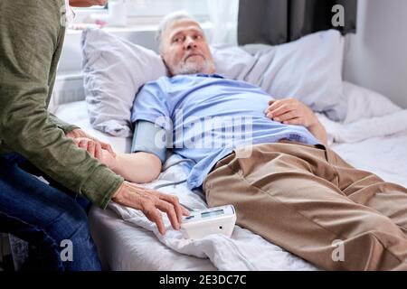 mature male patient at hospital with worried wife sitting with him, while checking blood pressure with tonometer. woman helps, support. focus on hands Stock Photo