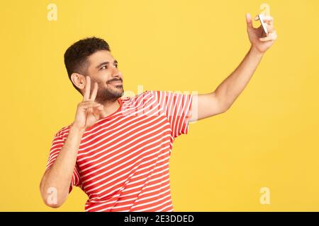 Cheerful popular man blogger in striped t-shirt showing victory gesture looking at smartphone camera, communicating with subscribers, streaming. Indoo Stock Photo