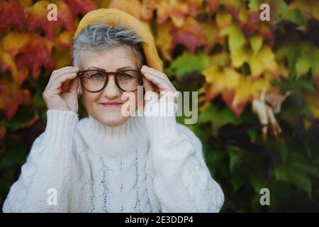 Senior woman standing outdoors against colorful natural autumn background, looking at camera. Stock Photo