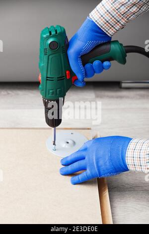 Electric Drill works in handyman hands at home. Male hands in gloves using electric drill tool to assemble and repair furniture. Home DIY Stock Photo