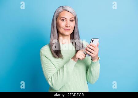Portrait of attractive satisfied elderly woman hold cellphone typing dressed green sweater isolated on blue color background Stock Photo