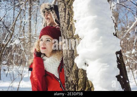 Playful couple hiding behind a tree trunk in the snow Stock Photo