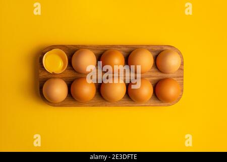 Wood tray with dozen farm brown chicken eggs with one yolk on yellow table. Minimalistic product concept. Easter holiday. Overhead view, flat lay Stock Photo