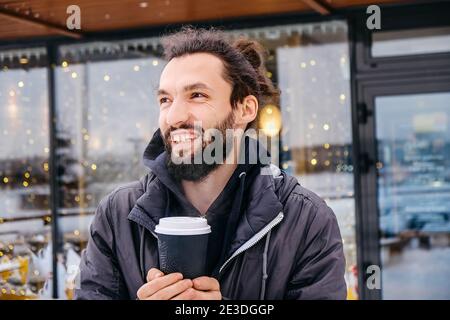 Portrait of cheerful attractive bearded young man drinking hot coffee outdoors in winter on cafe lights background Stock Photo