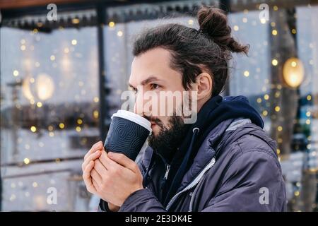 Portrait of attractive bearded young man drinking hot coffee outdoors. A man warms himself on hot coffee in winter. Take away service Stock Photo