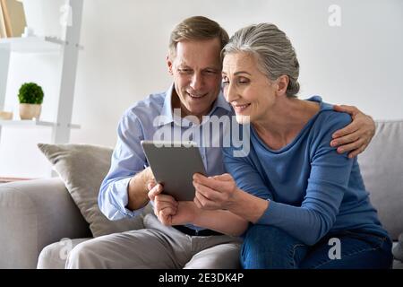 Happy old senior grandparents using digital tablet sitting on couch at home. Stock Photo