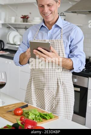 Old senior 50s man wearing apron using tablet preparing salad in kitchen. Stock Photo