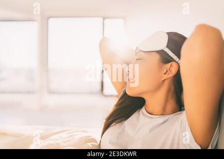 Sleeping at home woman waking up in morning sunlight stretching happy after a good night sleep feeling rested wearing eye mask. Asian girl bed sleep