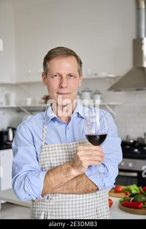 Confident middle aged old man holding wine glass standing in kitchen, portrait. Stock Photo