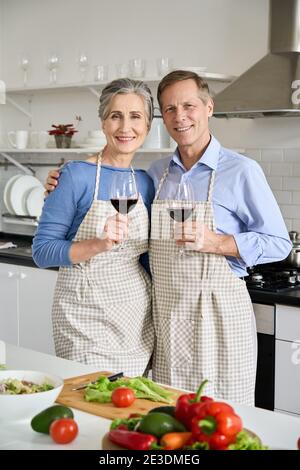 Happy old couple in aprons hugging, drinking wine, looking at camera in kitchen. Stock Photo