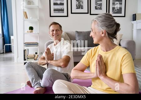 Happy healthy older senior 50s couple meditating with namaste hands at home. Stock Photo