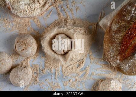 Flat lay image of bread loaves, dough ingredients, a bowl of flour, a sourdough bread on marble kitchen countertop. A dough loaf is dropped onto a pil Stock Photo