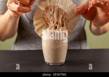 A woman is carefully removing the top cover of a sourdough starter culture which is about to overflow the glass cup. The gooey starter culture sticks Stock Photo