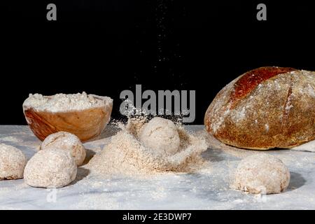 A bread loaf splash on a pile of wholewheat flour on marble countertop with dark background. There is fresh sourdough bread, a wooden bowl of flour an Stock Photo