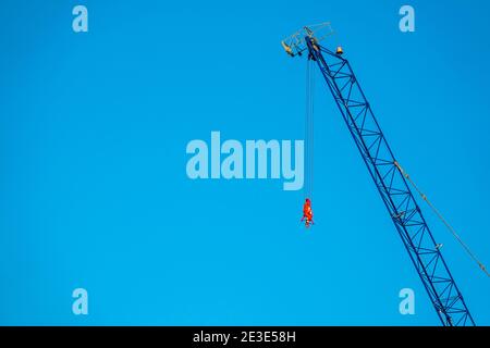Red lifting hook on a crane set against a blue sky above a construction site. Stock Photo