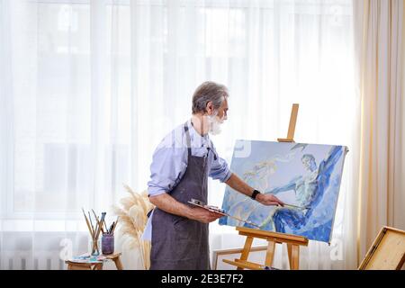painter creating masterpiece, senior male working on canvas, using paints, paintbrushes, easel and other tools Stock Photo