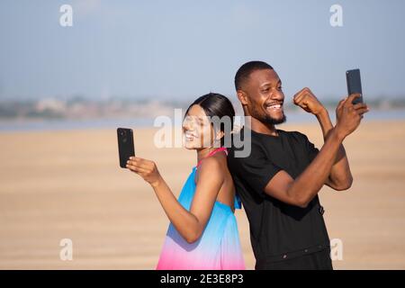 two young african friends standing back to back feeling excited and happy while looking at their phones Stock Photo