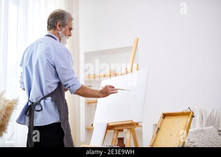 elderly artist man concentrated on painting, caucasian male in apron is immersed in the creation of art Stock Photo