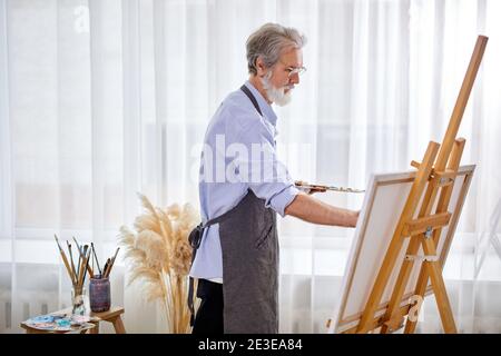 elderly artist man concentrated on painting, caucasian male in apron is immersed in the creation of art Stock Photo