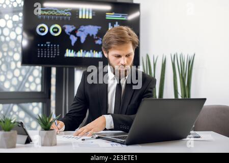 Horizontal indoor shot of young male concentrated financial director in formal wear, working at the desk in office, comparing datas from laptop with Stock Photo