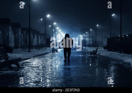 Silhouette of lonely person walks on dark foggy street illuminated with street lamps, blue toned. Stock Photo