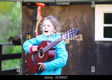 Little girl playing guitar and singing outdoors on green meadow at spring. Happy child on rural yard. Stock Photo