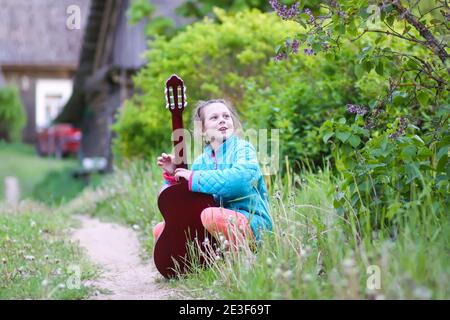 Little girl playing guitar and singing outdoors on green meadow at spring. Happy child on rural yard. Stock Photo
