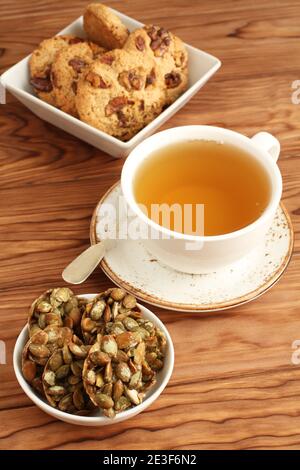 Oatmeal cookies with walnuts and raisins, sugar-coated pumpkin seeds cookies in a bowls and a cup of green tea on a wooden table. Closeup Stock Photo