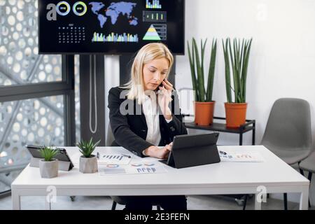 Horizontal shot of attractive focused high-skilled mature blond businesswoman which talking on phone and simultaneously typing on digital tablet Stock Photo