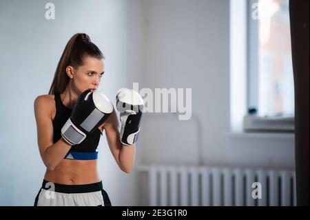 A pretty kickboxer girl is preparing for competitions in the boxing hall, practicing the technique of punches on a punching bag Stock Photo