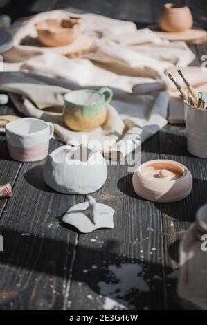 A close-up view of clay plates and a paint brush on a wooden table and dried flowers on a background. Crafted tableware in a pottery studio. Handmade Stock Photo