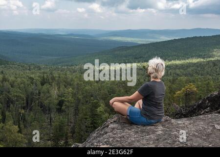A middle aged woman practices yoga and meditation on mountain top with stunning view of forested, hilly valley. Calm and relaxation. Healthy lifestyle Stock Photo