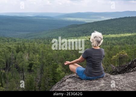 A middle aged woman practices yoga and meditation on mountain top with stunning view of forested, hilly valley. Calm and relaxation. Healthy lifestyle Stock Photo