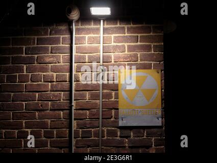 yellow and white fallout shelter sign on a brick wall at night with a spotlight shining on it creating a spooky, scary scene evoking danger Stock Photo