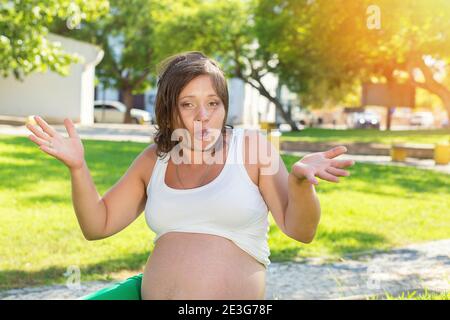 Emotional shrugging shoulders pregnant woman hands in air in showing I do not know gesture and I do not care with hands sitting outside, outdoors in a Stock Photo
