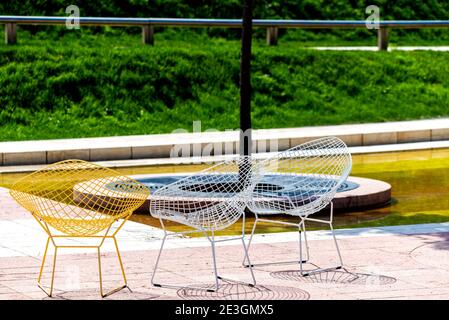 Wicker chairs made of wire stand near a tree on a tile. Nobody is there, unusual chairs are standing in a park. Stock Photo