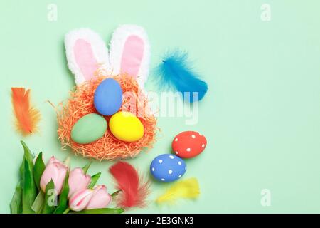 Easter eggs in a nest, bunny ears and flowers on a mint green background. Greeting card with copy space flat lay. Stock Photo
