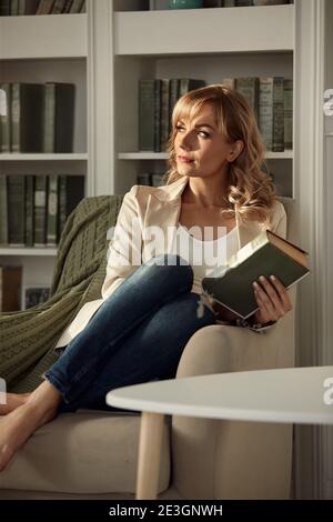 Young woman chilling at home. Girl relaxes and reads a book in the rays of the cozy warm sun. Sitting on retro chair near the window in light her livi Stock Photo