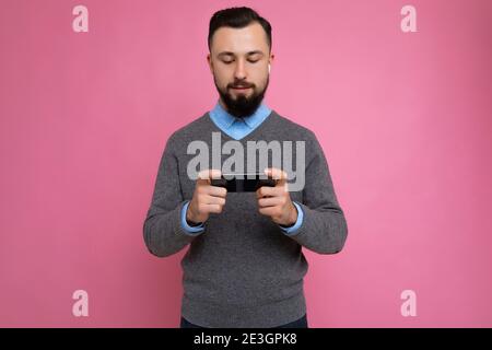 Photo shot of cool good looking brunet unshaven young man with beard wearing grey sweater and blue shirt isolated over pink background wall with empty Stock Photo