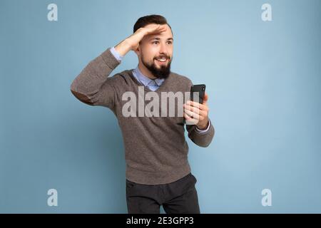 closeup portrait of Handsome smiling brunette bearded man wearing grey sweater and blue shirt isolated on background wall holding smartphone looking Stock Photo