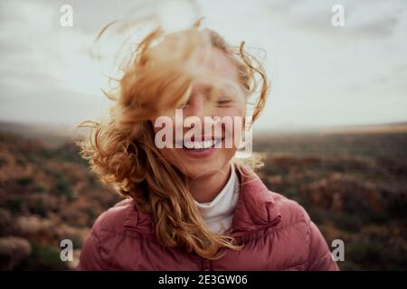 Portrait of young smiling woman face partially covered with flying hair in windy day standing at mountain - carefree woman Stock Photo