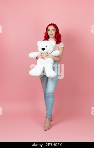 full-length beautiful, delicate woman in jeans with red hair hugs a large  white teddy bear isolated on a pink background Stock Photo - Alamy