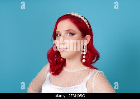 close-up face of a smiling young woman with red hair in a white dress, pearl headband and long dangling earrings, isolated on a blue background Stock Photo