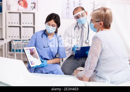 Doctor exmplaining heart disease to senior woman at hospital cardiology wearing face mask using tablet pc. Medical assistant having a conversation with elder patient during coronavirus outbreak. Stock Photo