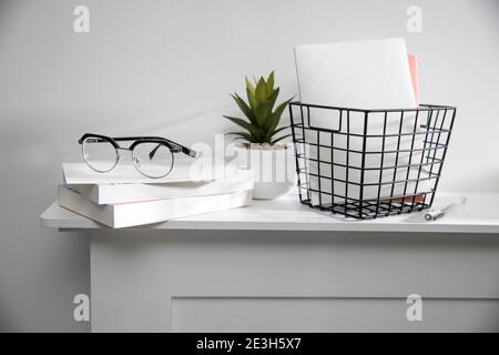 A stack of books, an artificial succulent plant in a ceramic pot, glasses, a metal basket with notebooks, a pen and a cup on a white table surface. Co Stock Photo