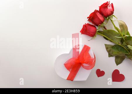 Romantic background with three red roses and a white gift box with a red heart-shaped bow and cutouts of red hearts on a white table. Top view. Stock Photo